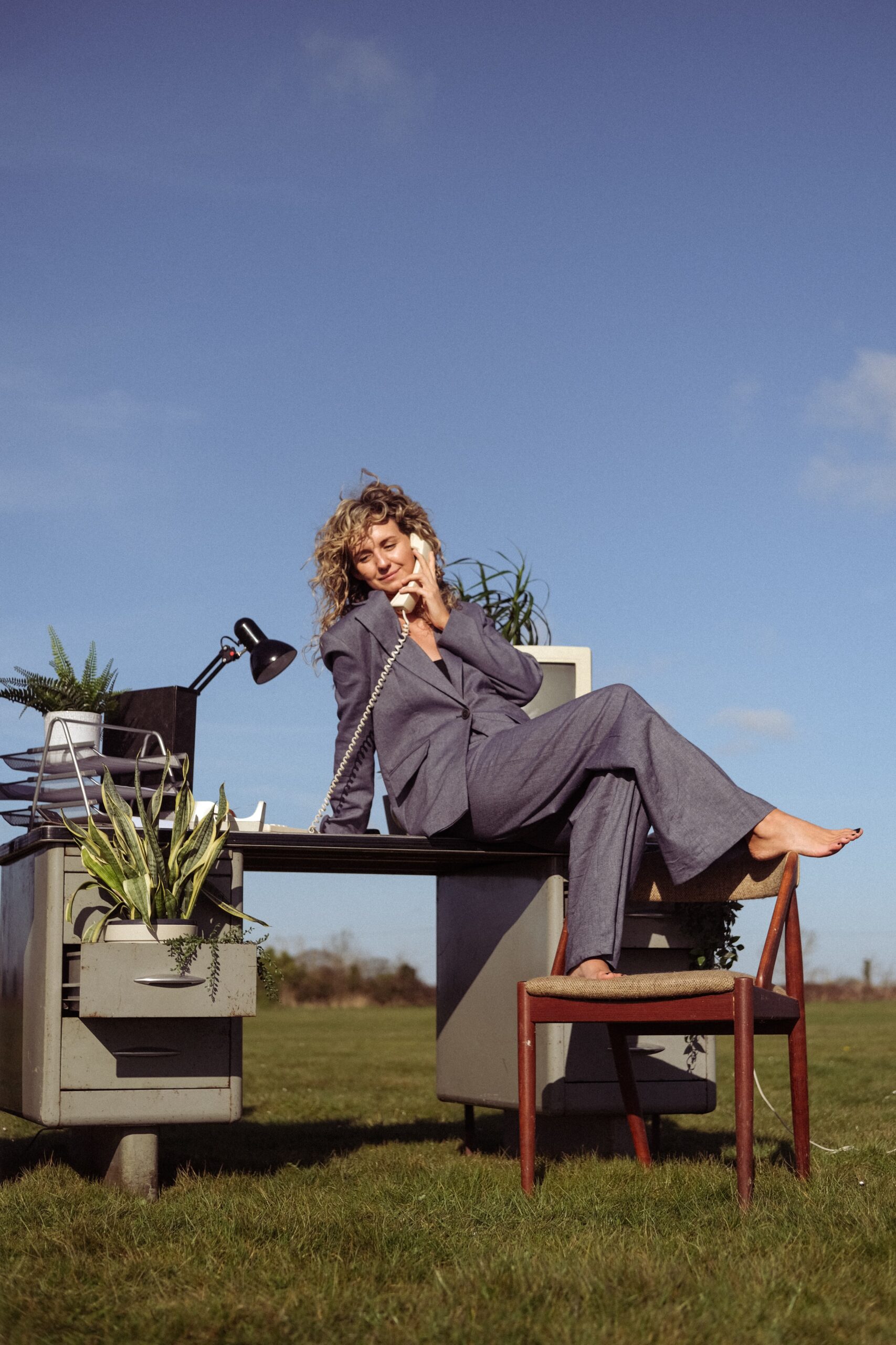 Woman in a grey suit casually sitting on an outdoor desk, using an old landline phone. Plants and office supplies surround her, set against a clear blue sky.