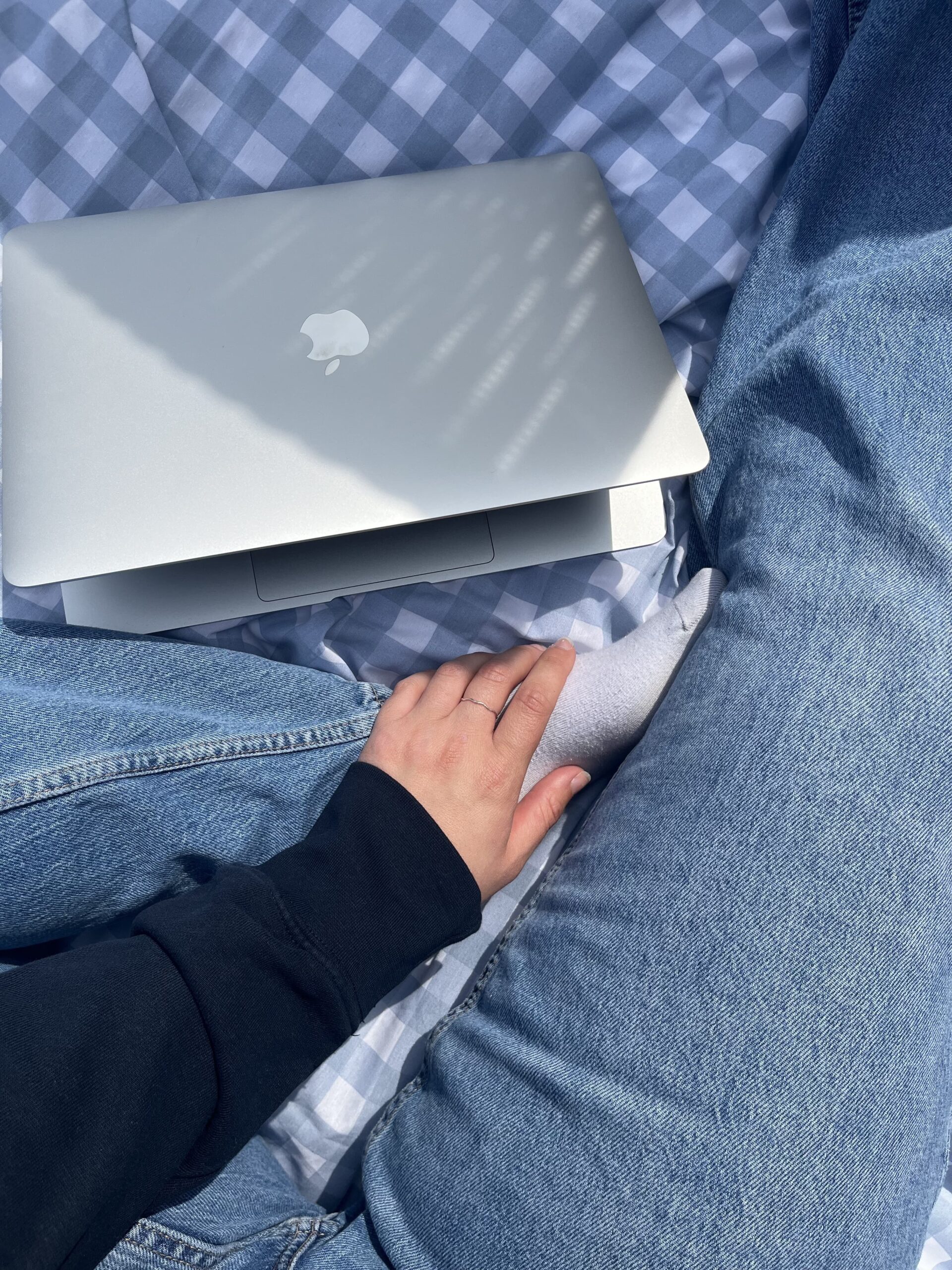 Close-up overhead image of a person wearing jeans sitting on a light blue checkered blanket with a silver laptop. Their hand is gently placed on their folded left leg.