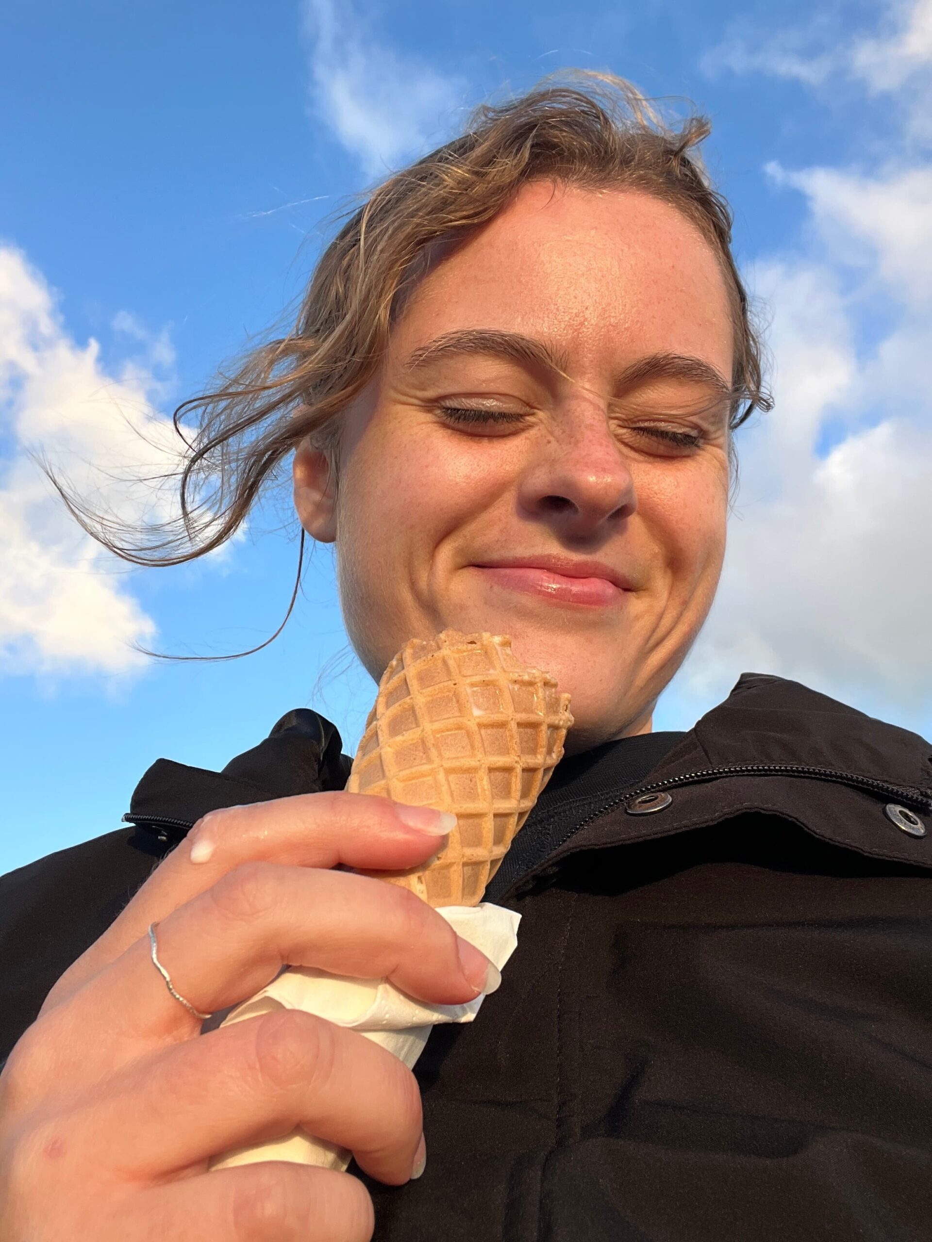 Olivia smiling with closed eyes holding an ice cream cone under a clear blue sky with a few clouds. Wearing a black coat.
